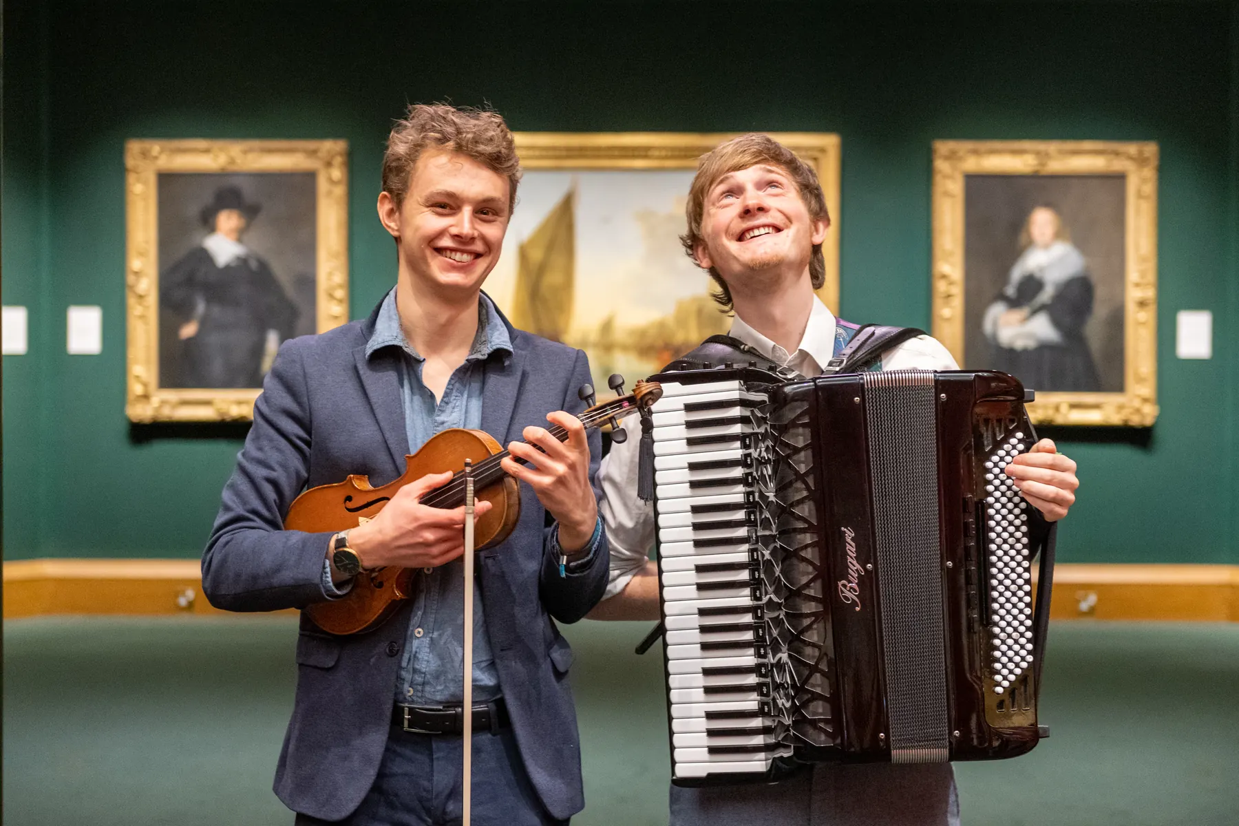 Roo playing fiddle and Neil holding his accordion, smiling in front of framed paintings