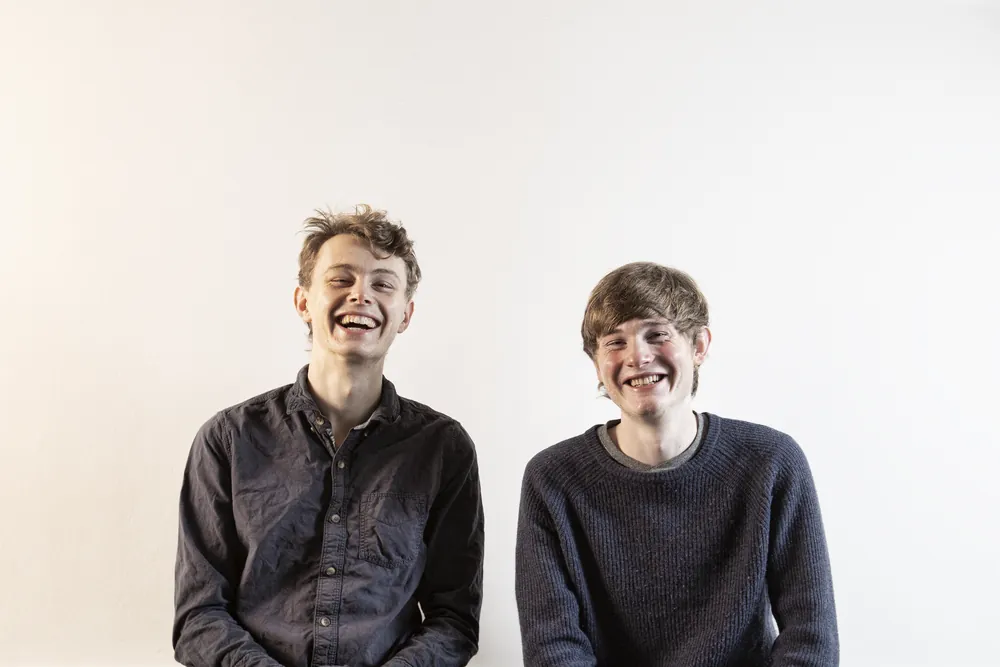 Roo and Neil laughing together against a white studio wall